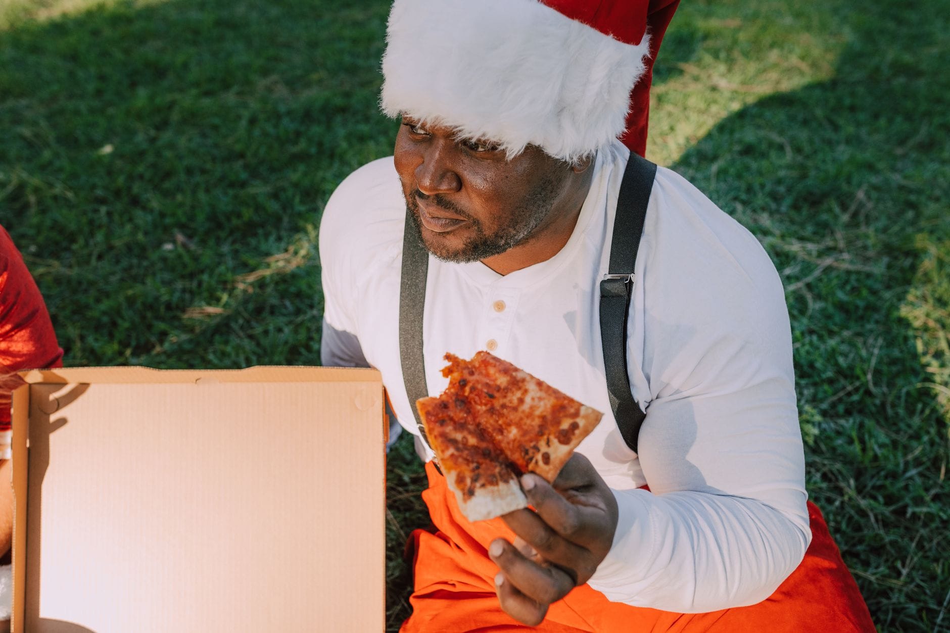 Mężczyzna w stroju świętego Mikołaja je pizzę na pikniku na świeżym powietrzu Mężczyzna w stroju świętego Mikołaja je pizzę na pikniku na świeżym powietrzu